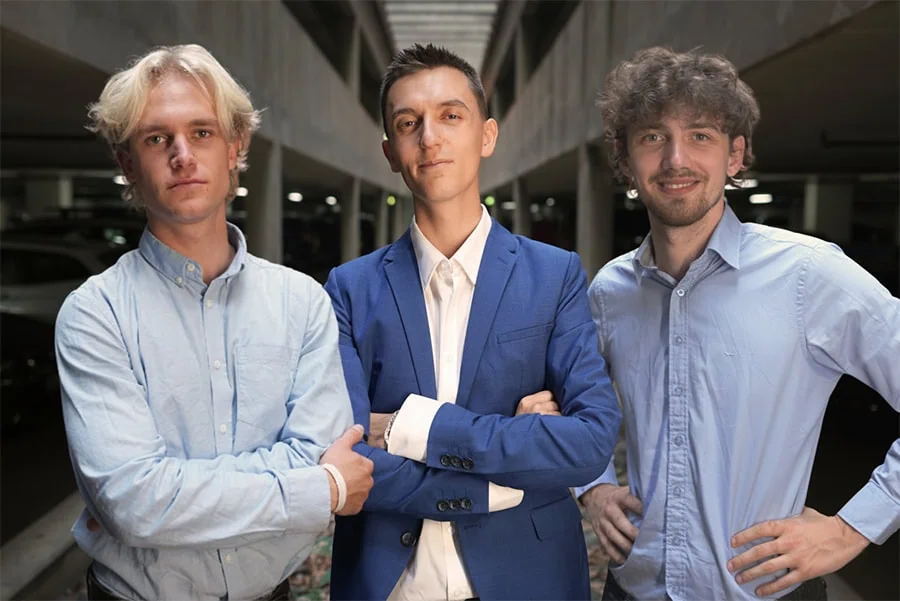 Drei junge Männer stehen in einer Parkgarage. Der Mann in der Mitte trägt einen blauen Anzug, während die anderen beiden Hemden tragen.